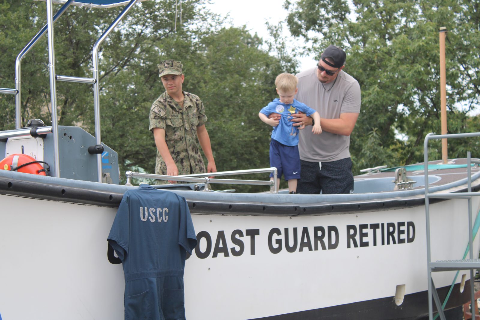 A Sea Cadet and a father lifting a toddler aboard the retired Coast Guard surf boat RES 1 at a community event